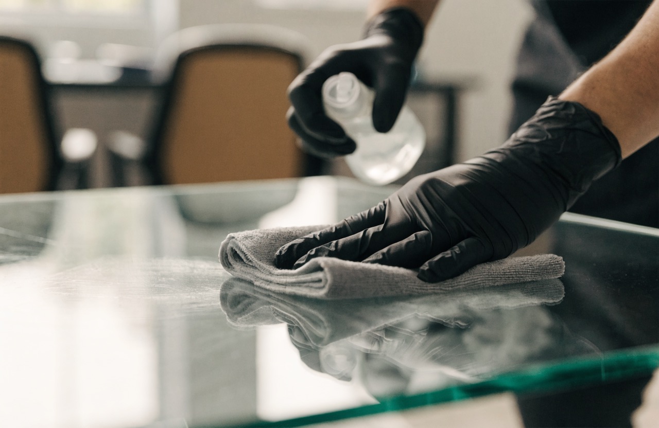 Reinigungsfachkraft desinfiziert mit Handschuhen, Sprühflasche und Mikrofasertuch eine Glasoberfläche
